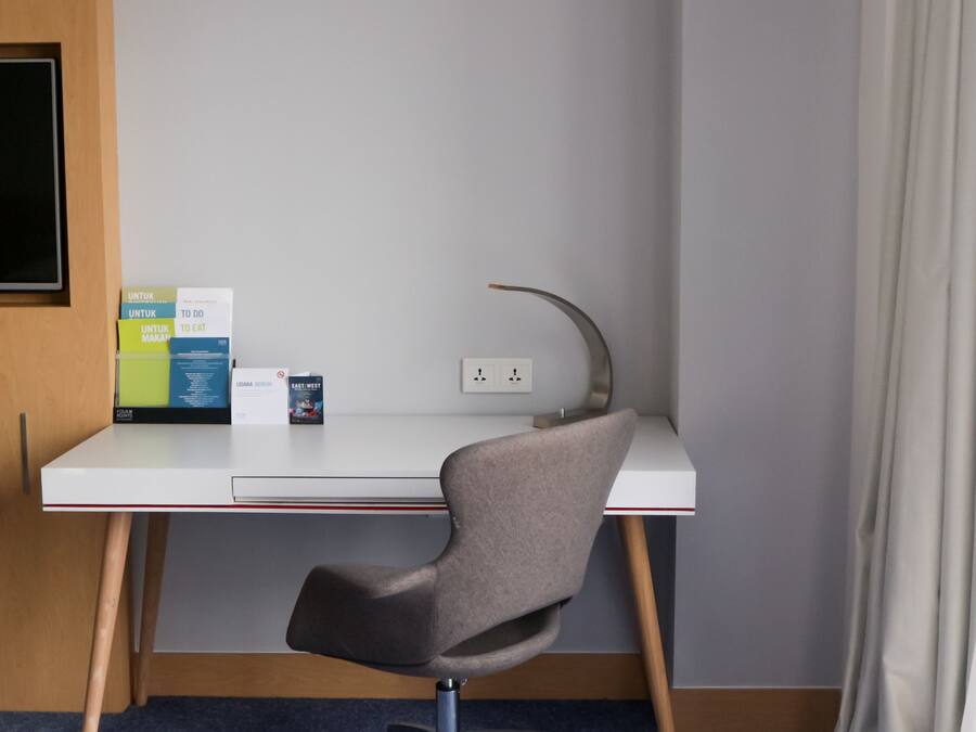 A modern hotel room with a white desk, a grey chair, and a small bookshelf with books and a magazine.