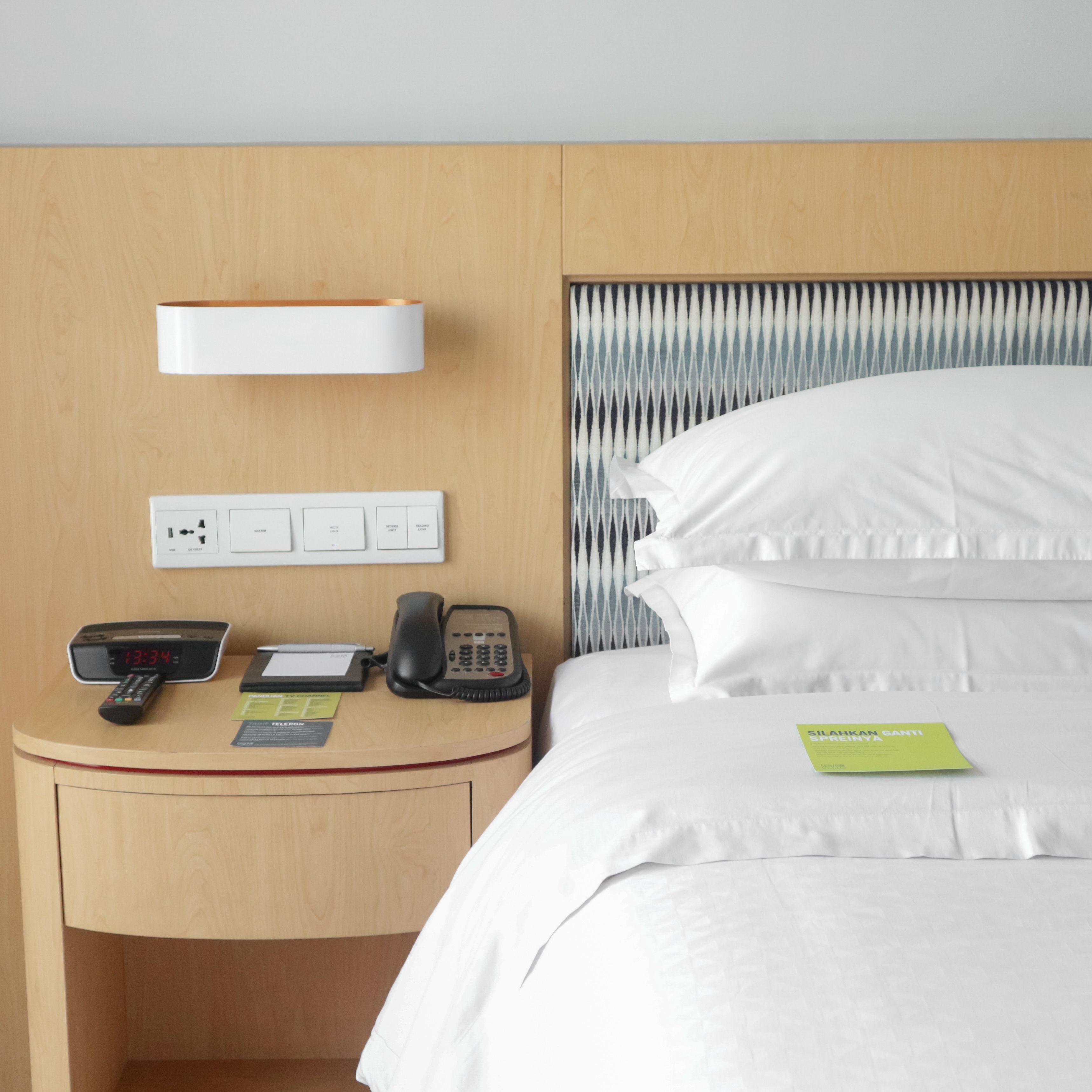 Una habitación de hotel con una mesita de noche de madera, una cama con ropa de cama blanca, un teléfono, un reloj digital y una tarjeta pequeña sobre la cama.