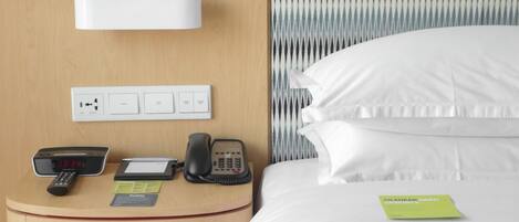 Una habitación de hotel con una mesita de noche de madera, una cama con ropa de cama blanca, un teléfono, un reloj digital y una tarjeta pequeña sobre la cama.