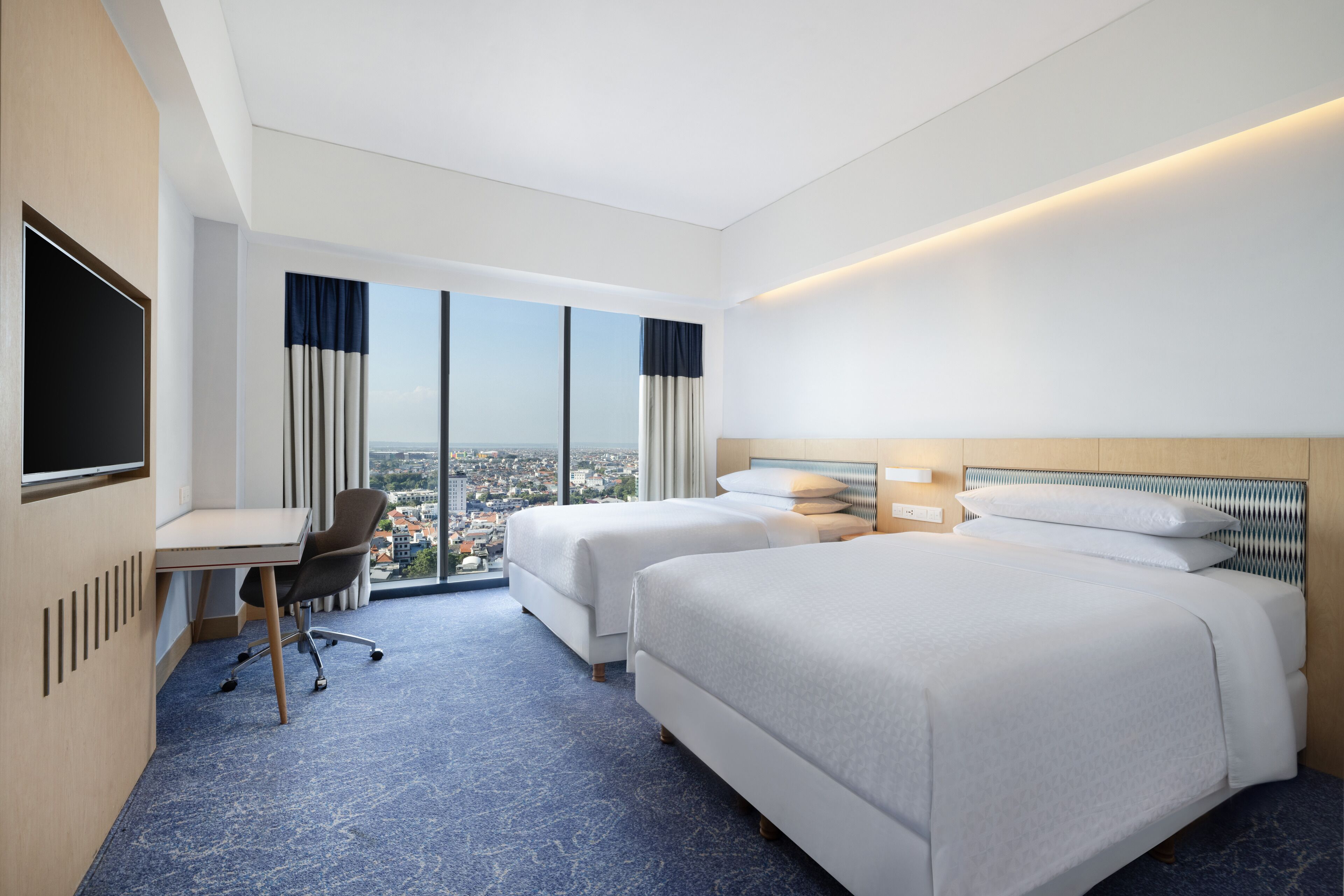 Habitación de hotel con dos camas, un escritorio y un amplio ventanal con vistas a la ciudad.