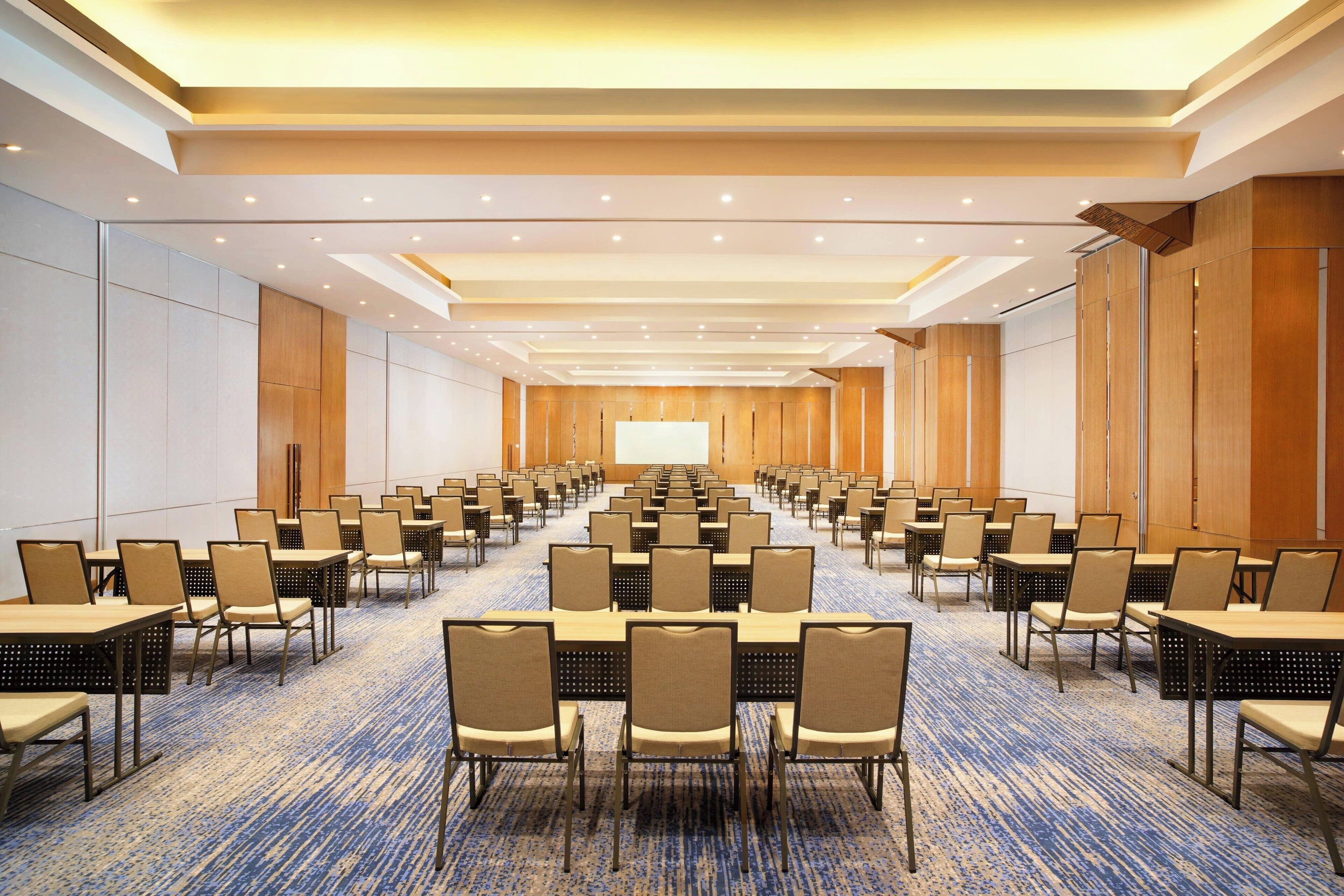 A conference room with rows of tables and chairs, a large screen at the front, and a carpeted floor.