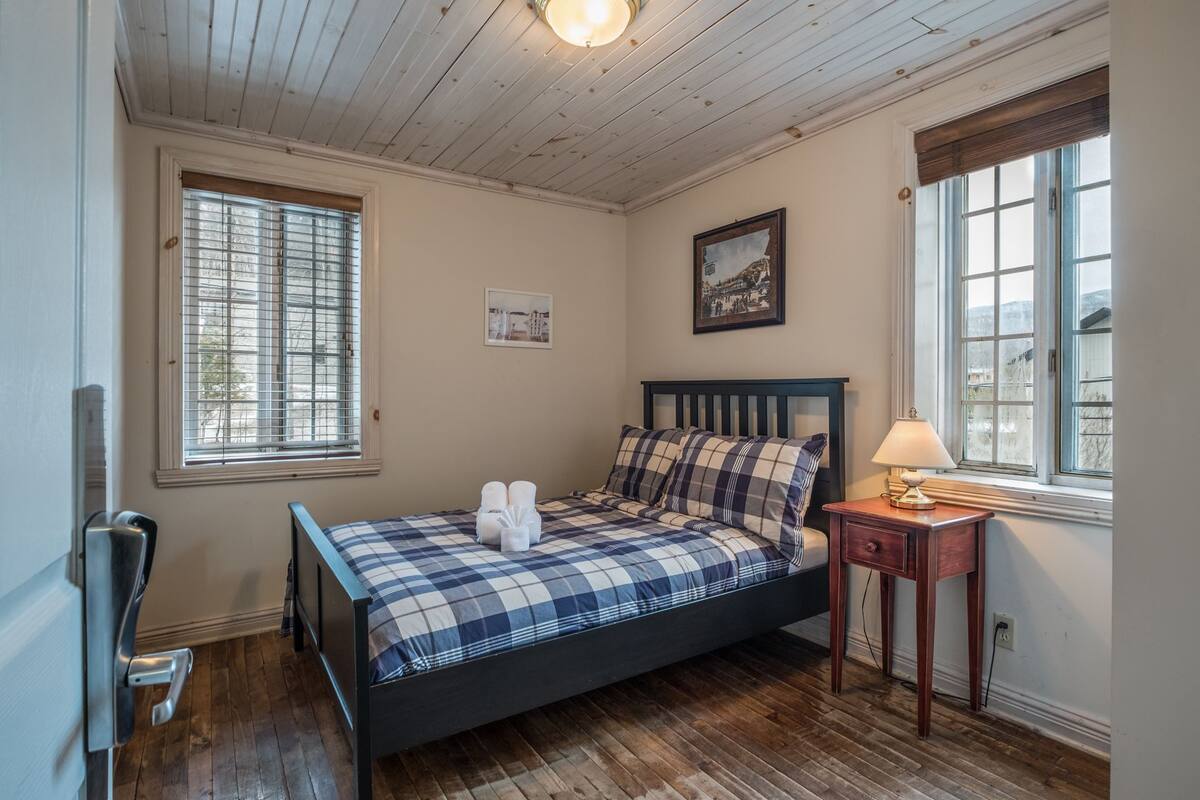 A bedroom with a bed, a bedside table, a lamp, and two windows.
