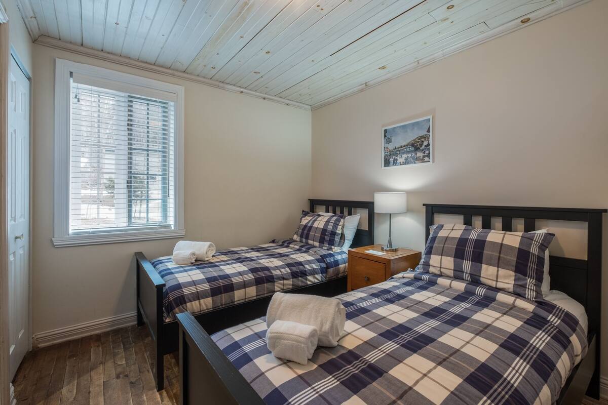 A bedroom with two beds, a window with blinds, a nightstand with a lamp, and a painting on the wall.