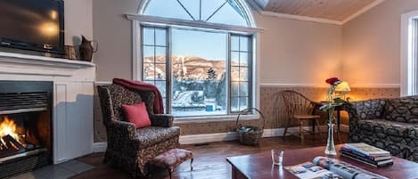 A cozy living room with a fireplace, a large window with a view, a patterned armchair, a sofa, and a coffee table with magazines.