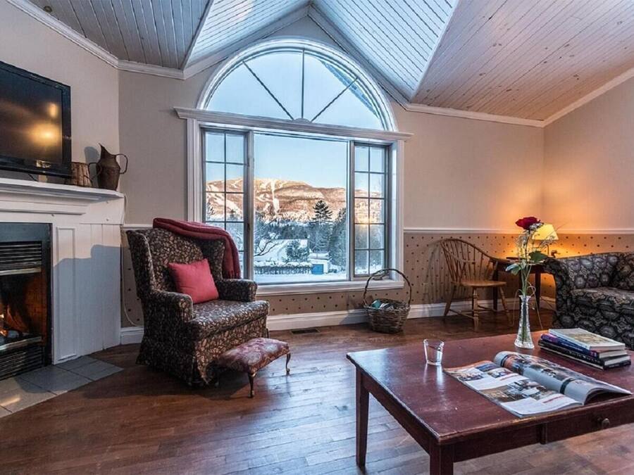 A cozy living room with a fireplace, a large window with a view, a patterned armchair, a sofa, and a coffee table with magazines.