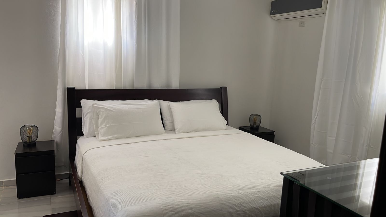 Un lit bien fait, avec du linge de lit blanc, deux tables de chevet avec des lampes et un climatiseur fixé au mur.