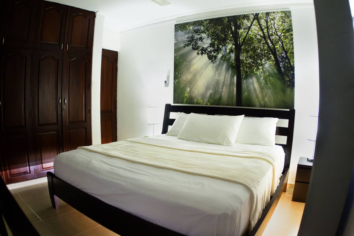 Ein ordentlich bezogenes Bett mit weißen Leinentüchern in einem Zimmer mit großem Fenster, einem dunklen Holzschrank und einem Wandbild eines Waldes.