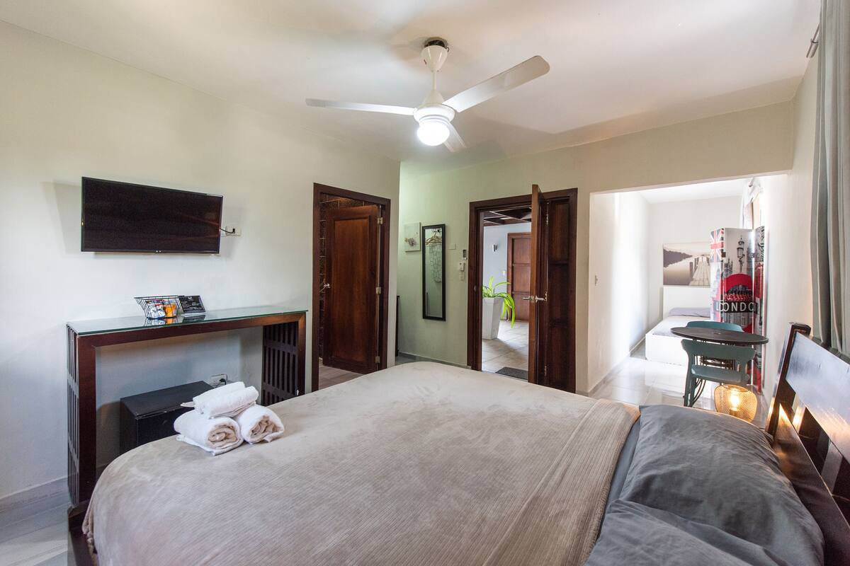 Ein Hotelzimmer mit Bett, Schreibtisch, Deckenventilator und an der Wand montiertem Fernseher.