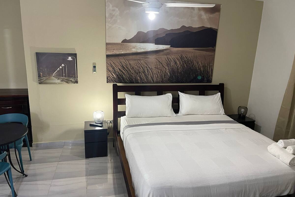 Ein Hotelzimmer mit Bett, Schreibtisch mit Stuhl, Nachttisch und Deckenventilator.