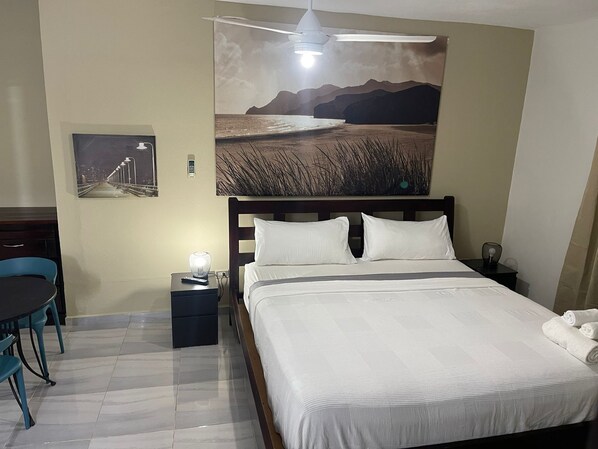 A hotel room with a bed, a desk with a chair, a nightstand, and a wall-mounted ceiling fan. - Hotel Casa Jum (Santiago de los Caballeros)