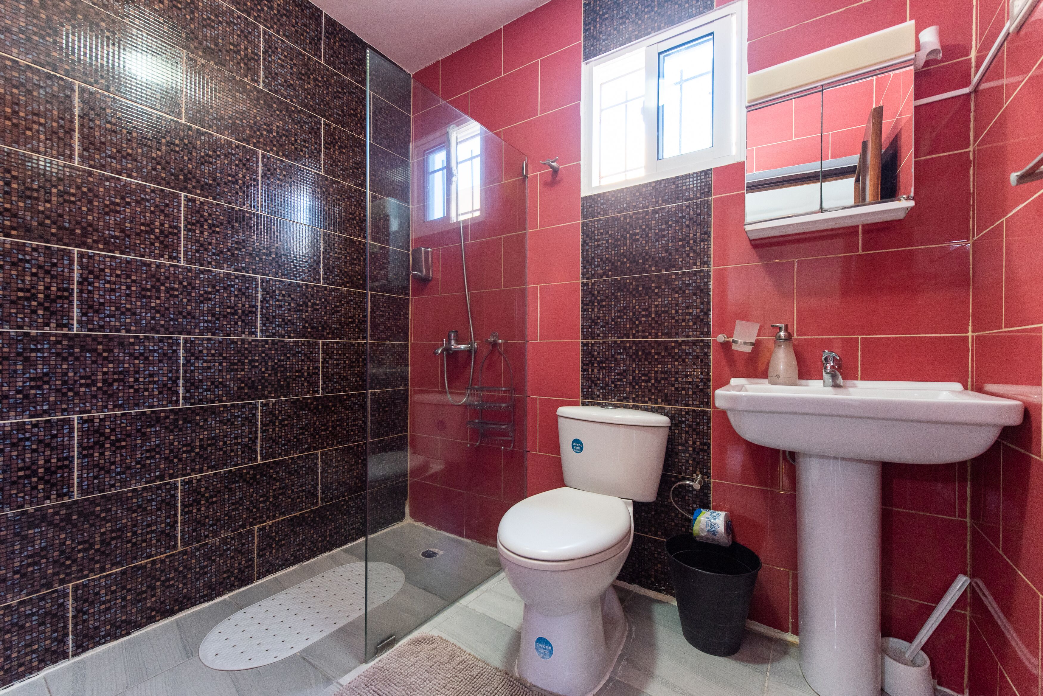 A bathroom with a glass shower enclosure, a white toilet, a sink, and a red tiled wall.
