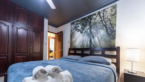 A bedroom with a large bed, a nightstand, a lamp, and a painting of a forest on the wall. - Hotel Casa Jum (Santiago de los Caballeros)