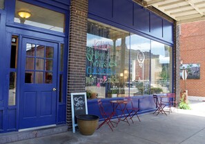 A storefront with a blue door and large windows displaying a chalkboard sign advertising 'HUMM'S WRAPS COLD BREW mint-rose matcha'. There are outdoor tables and chairs, a chalkboard sign, and a trash can outside.
