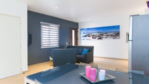 Een moderne woonkamer met een glazen tafel, een bank, een schilderij van een strand en een koelkast.