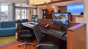 Uma área de recepção de hotel com duas estações de trabalho de computador, um espaço de estar com um sofá azul e um balcão de recepção com monitor de computador exibindo uma imagem do resort.