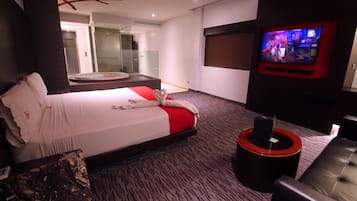Habitación de hotel moderna con una cama grande, televisor, zona de estar y baño visible a través de una puerta de vidrio.