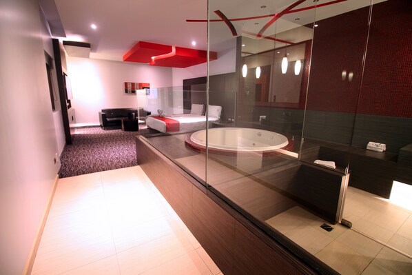 Quarto de hotel moderno com hidromassagem em vidro, cama com parede de destaque vermelha e área de estar separada com sofá e mesa de centro.