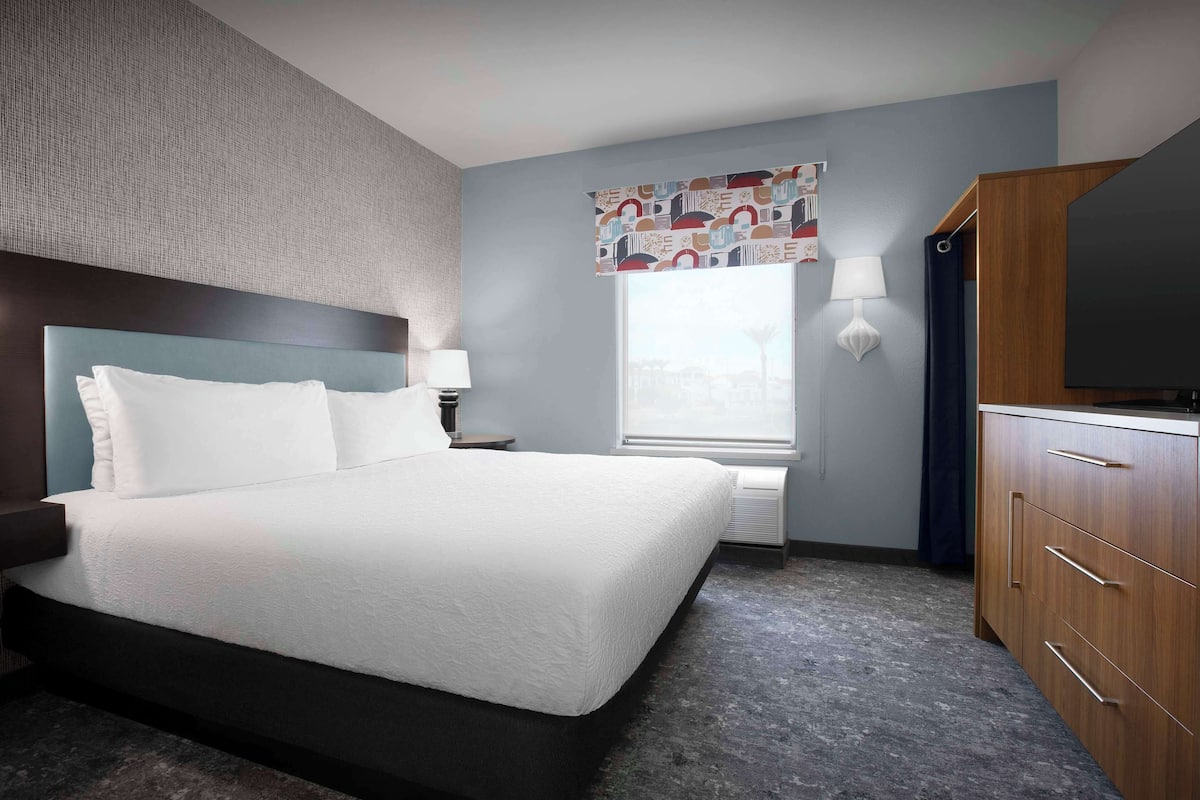 A hotel room with a large bed, a wooden dresser, a window with a patterned curtain, and a wall-mounted TV.