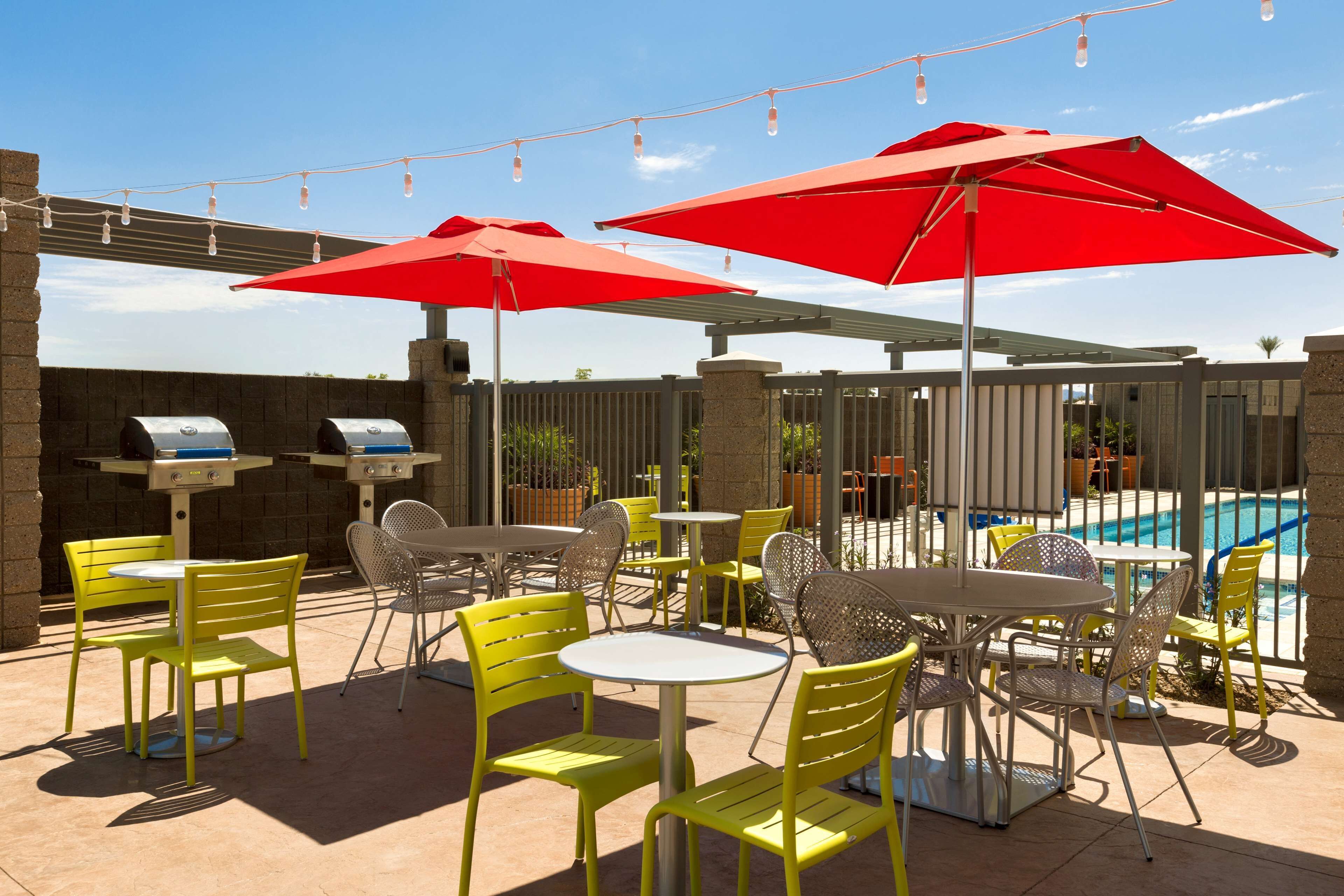 An outdoor patio with yellow chairs and tables, red umbrellas, and a pool in the background.