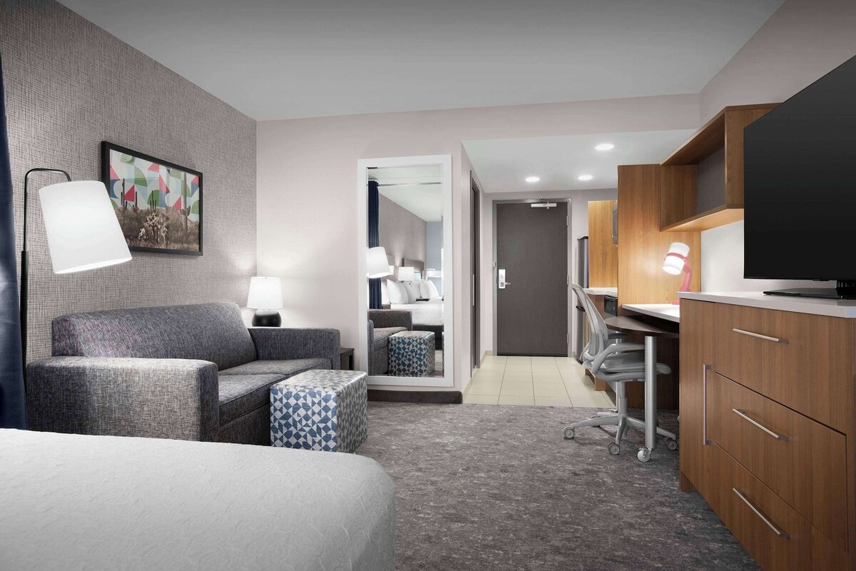A hotel room with a bed, a sofa, a desk with a chair, a TV, and a mirror.