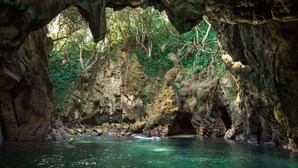 Une grotte où coule une rivière, entourée de falaises rocheuses et de verdure.