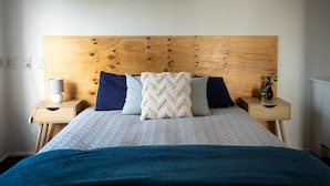 Une chambre à coucher avec une tête de lit en bois, un lit recouvert d’une courtepointe blanche et un tableau représentant une vague accroché au mur.