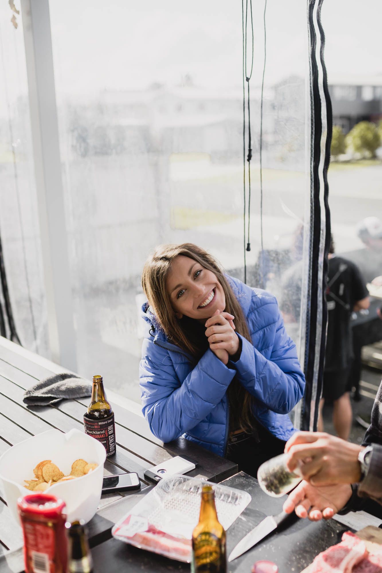 A person sitting at a table with a bottle of beer, a bowl of chips, and a bottle of sauce.