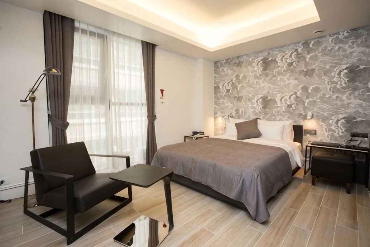 A modern hotel room with a bed, a desk, a chair, and a nightstand.