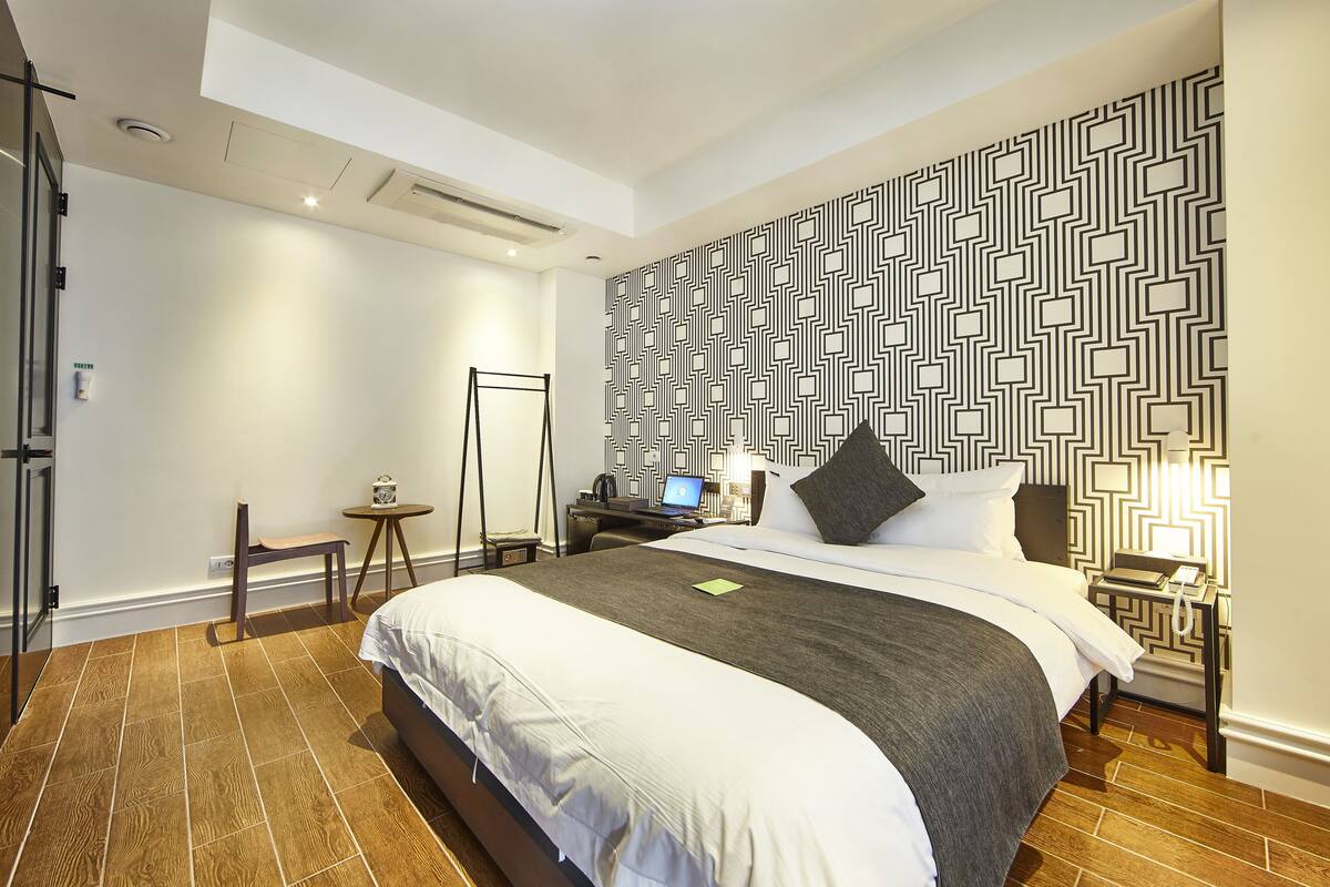 A hotel room with a large bed, a desk with a laptop, a chair, and a patterned wall.