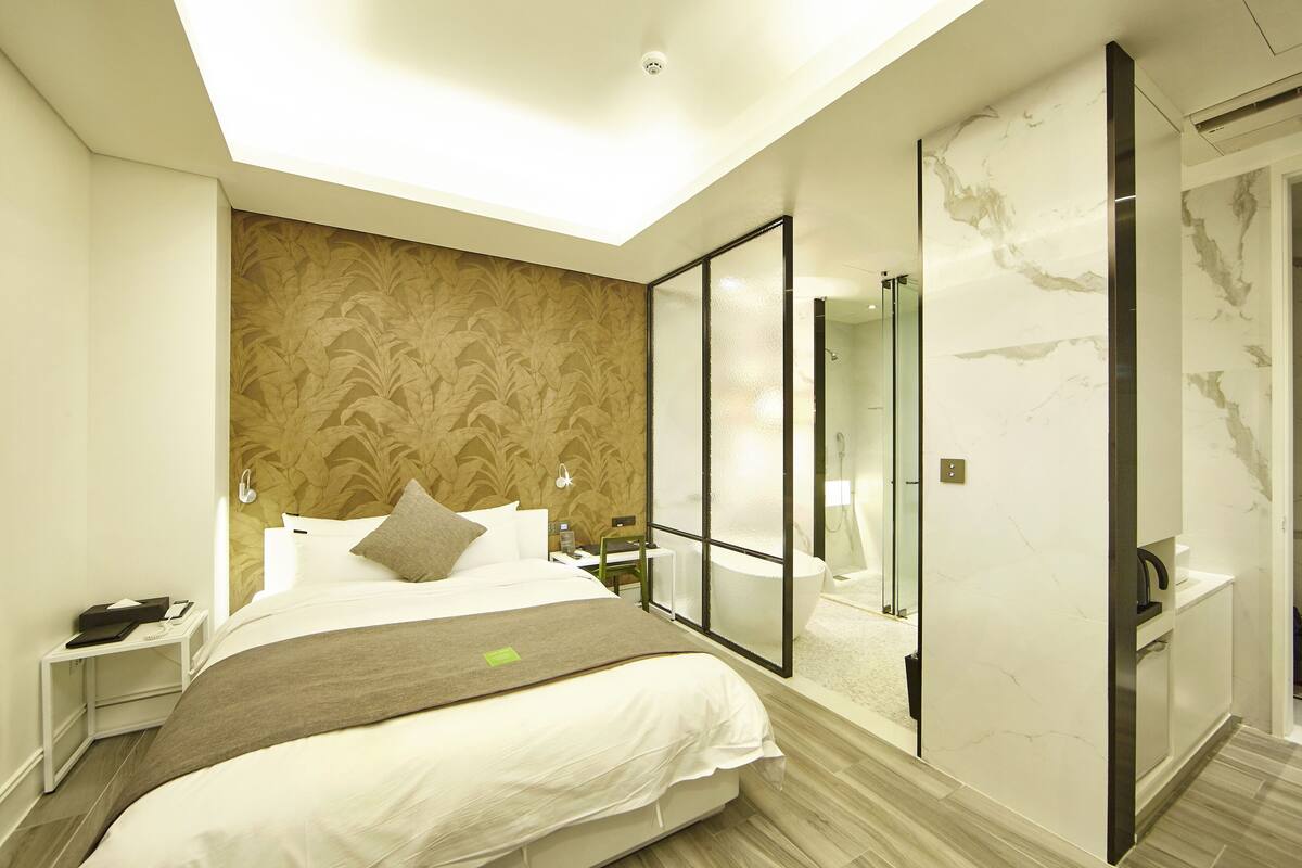 A modern hotel room with a large bed, a glass partition, a bathroom with a marble wall, and a ceiling light.