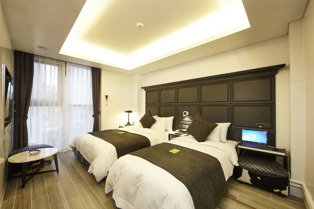 A hotel room with two beds, a desk with a laptop, a small table, and a TV.