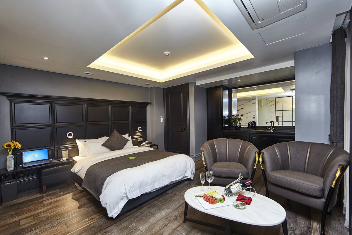 A modern hotel room with a large bed, a desk with a laptop, two armchairs, a dining table with a plate of food and wine glasses, and a large mirror.