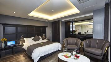 Una habitación de hotel moderna con una cama grande, un escritorio con una computadora portátil, dos sillones, una mesa de comedor con un plato de comida y copas de vino, y un espejo grande.