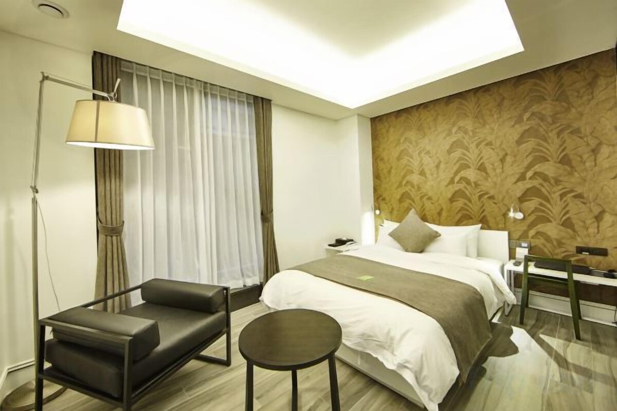 A hotel room with a bed, a desk, a chair, a lamp, and a window with curtains.