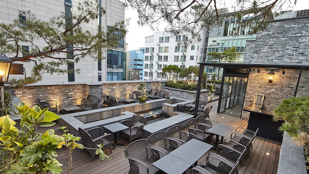 An outdoor patio with multiple seating areas, including tables and chairs, surrounded by greenery and modern buildings in the background. - Hotel Soulhada (Seoul)