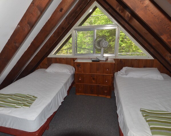 A small attic bedroom with two single beds, a wooden dresser, and a window with a fan. - Balsam Resort (Kawartha Lakes)