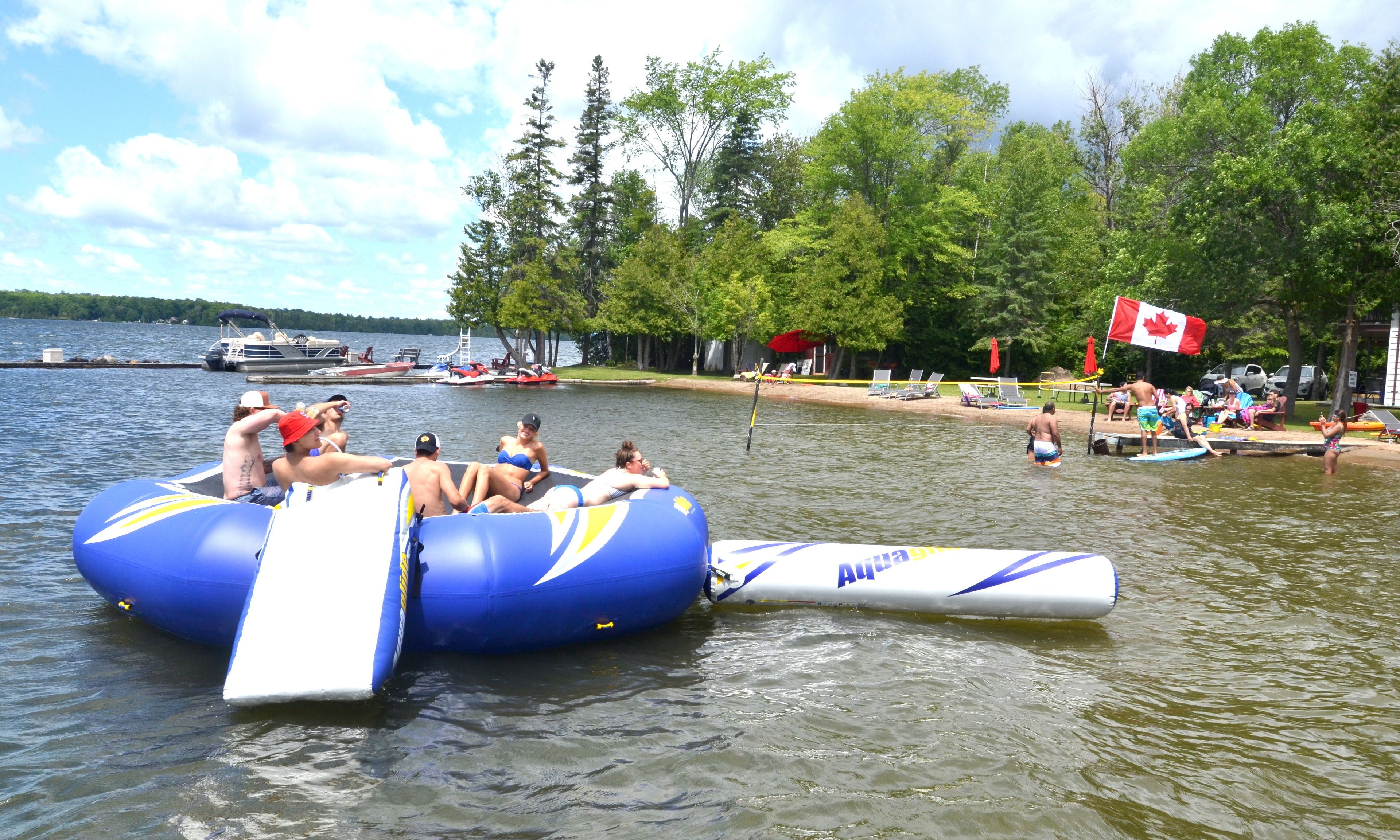 A group of people on a blue inflatable raft with a Canadian flag, floating on a lake with boats and a dock in the background.