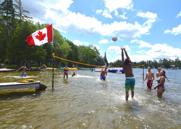 A lake with people playing volleyball, a Canadian flag, and a dock with boats. - Balsam Resort (Kawartha Lakes)