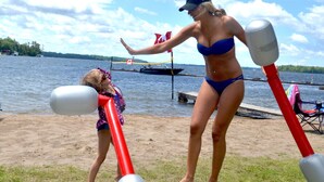 Une personne vêtue d’un bikini bleu et d’une casquette, debout sur une plage de sable, avec un lac en arrière-plan.