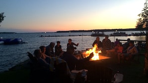 Un groupe de personnes s’est rassemblé autour d’un feu de camp au bord d’un lac, à la tombée de la nuit.