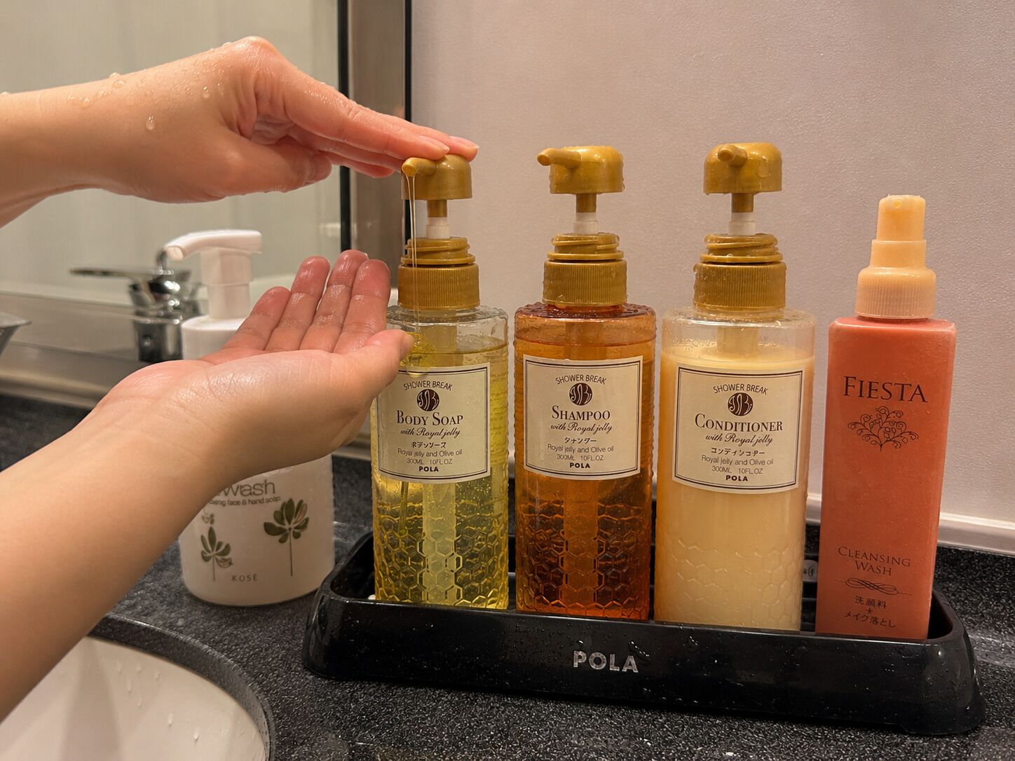 A hand selecting a bottle from a set of toiletries on a black tray.