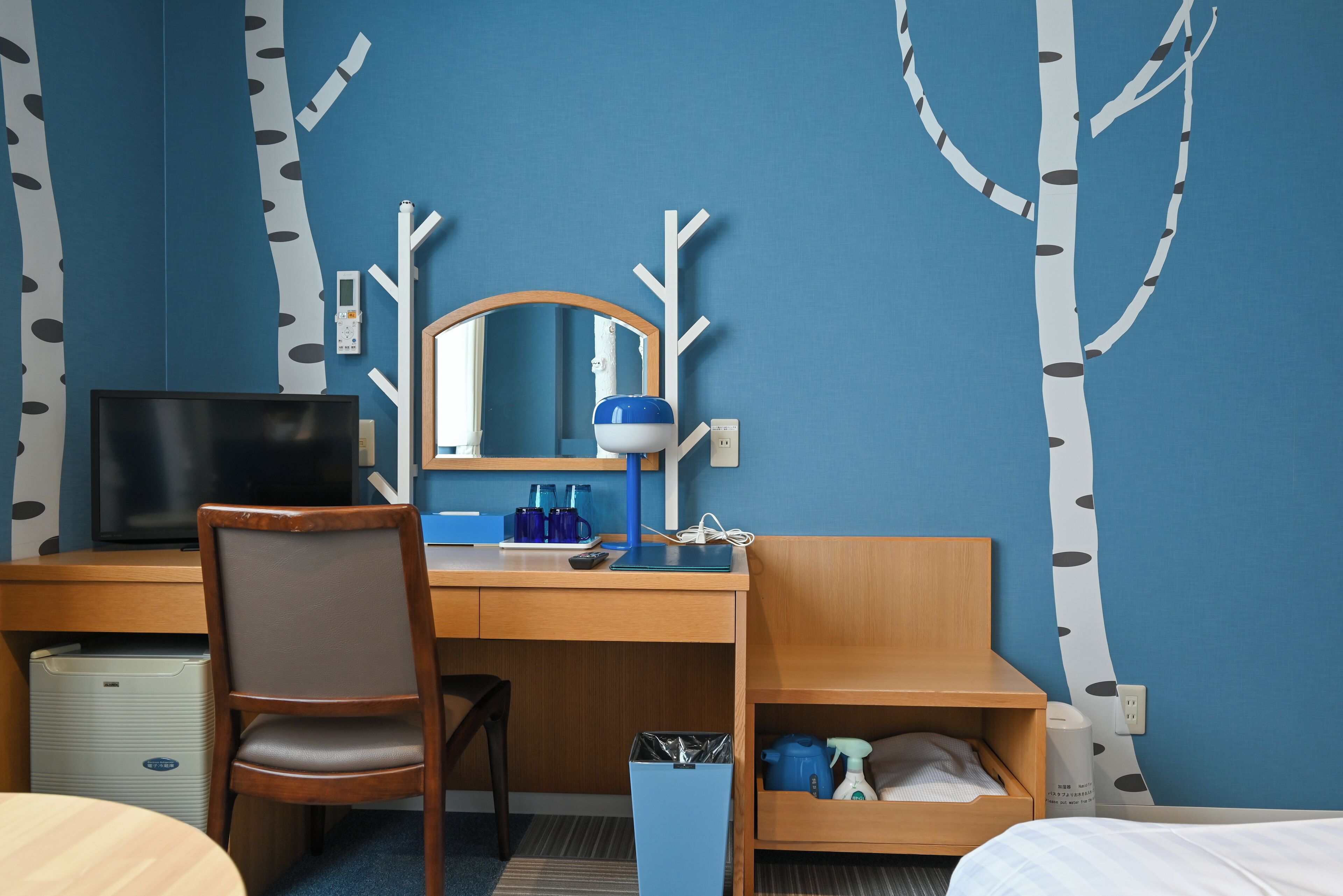 Habitación de hotel con un escritorio de madera, una silla, un televisor, un espejo, un estante con artículos de aseo y una pared con estampado de árbol.