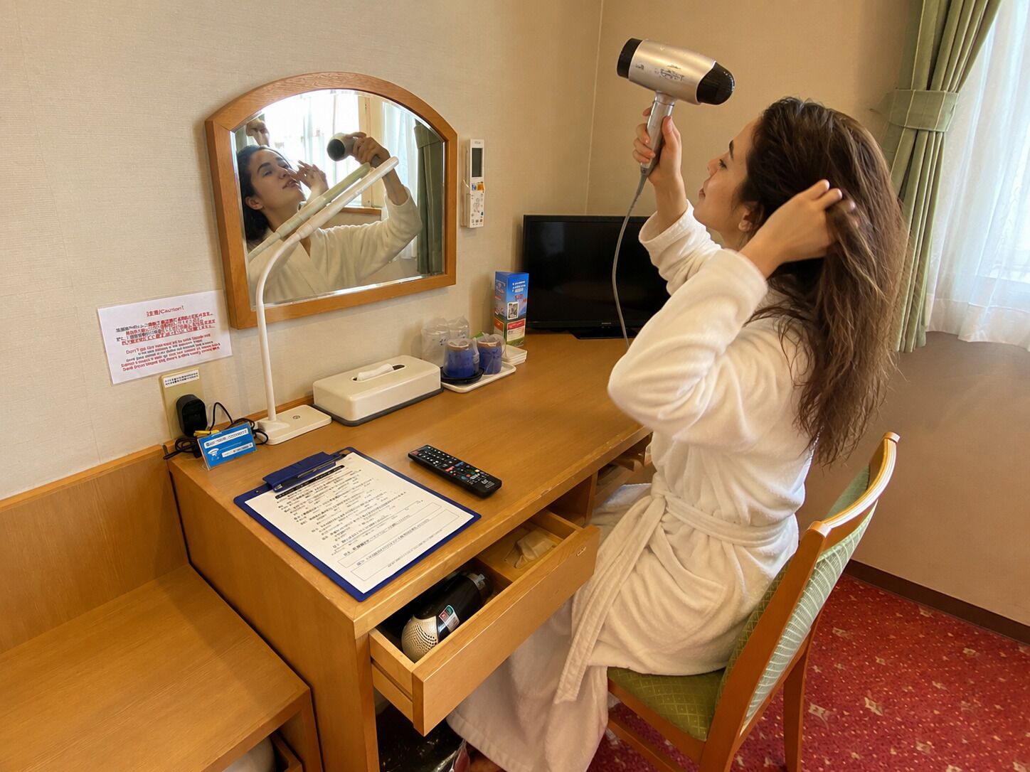 A hotel room with a wooden desk, a mirror, a hair dryer, a remote control, a tissue box, and a sign with safety instructions.
