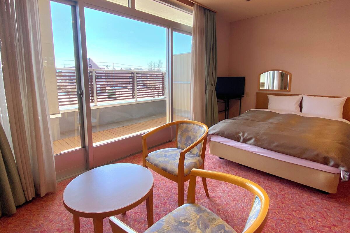 A hotel room with a bed, a small table, two chairs, a TV, and a balcony with a view of buildings.