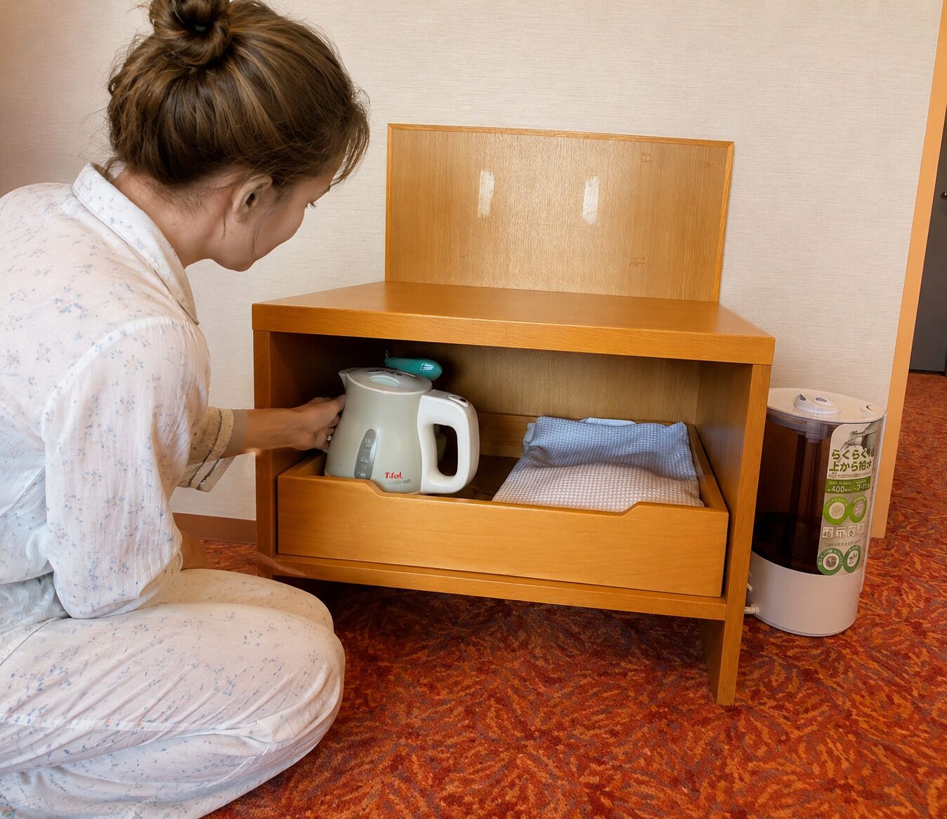 A person in pajamas opening a wooden drawer unit with a kettle and a small box inside.