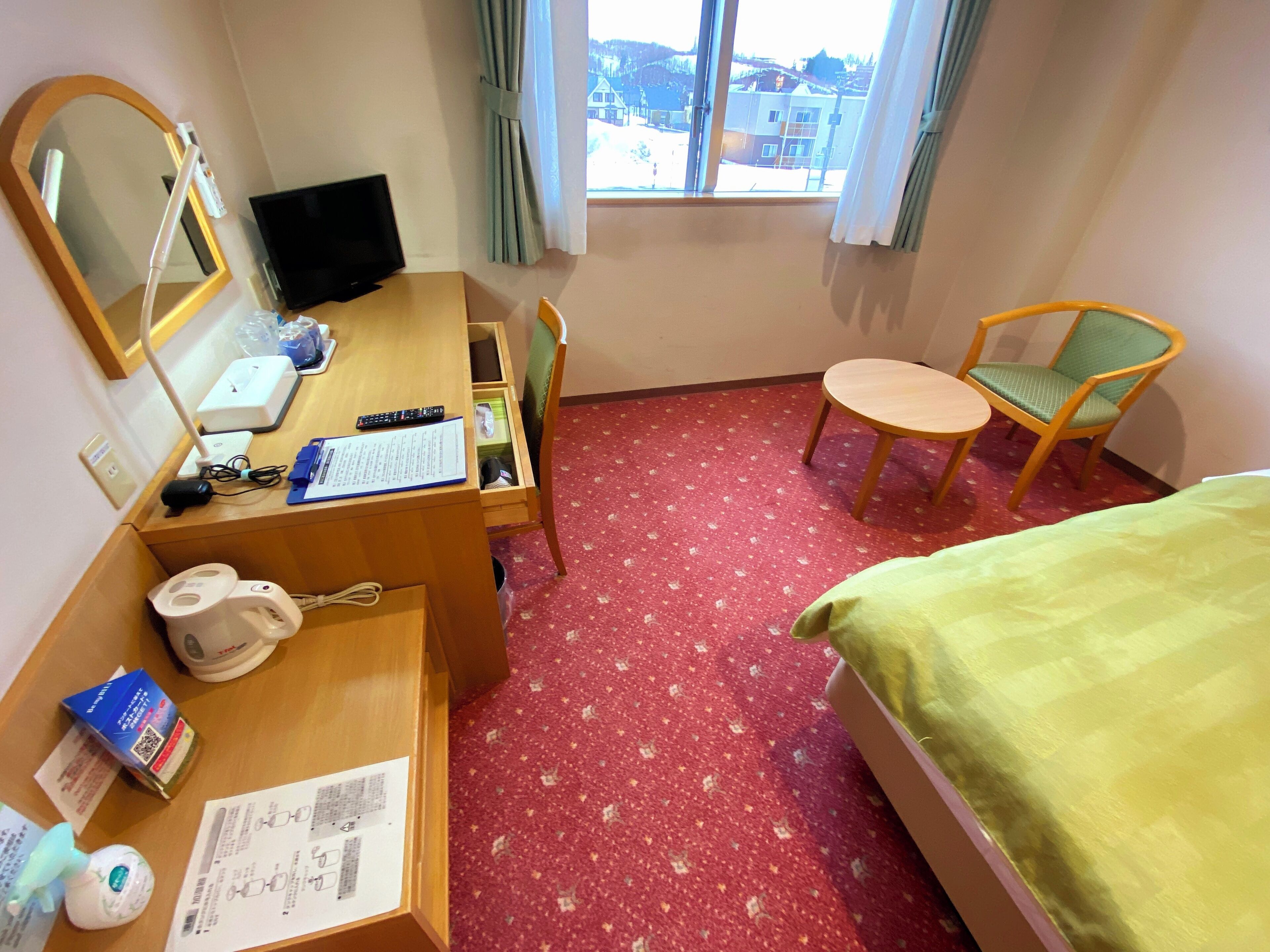 A hotel room with a bed, a desk with a computer, a chair, a window with curtains, and a red carpet.