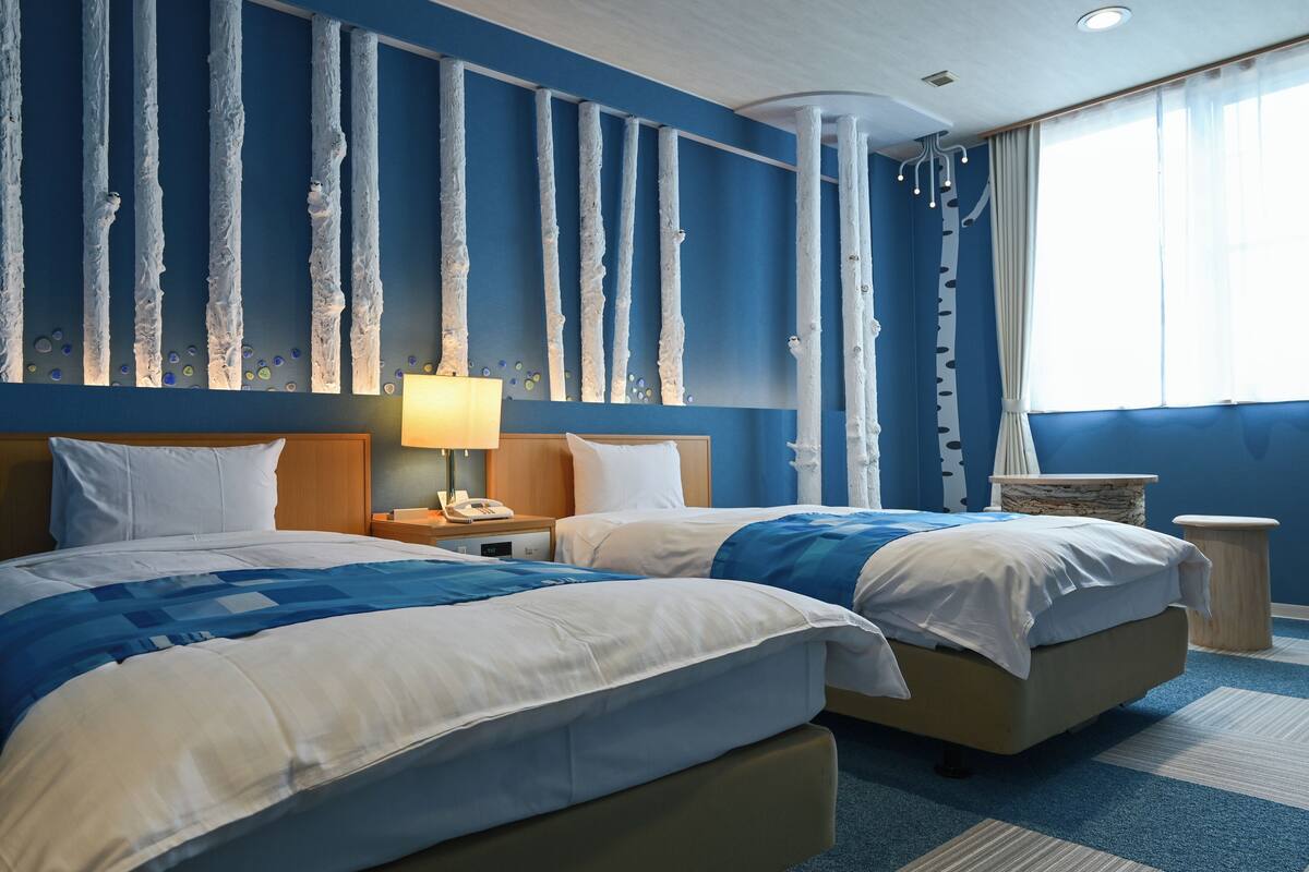 A hotel room with two beds, a nightstand, a lamp, and a large window with curtains.
