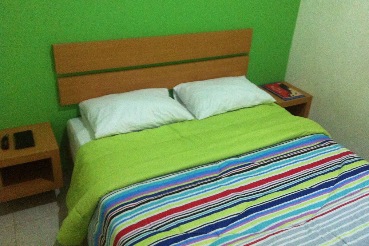 Uma cama com cabeceira verde, almofadas brancas e um cobertor listrado e colorido. Há duas mesas de cabeceira de madeira, com um livro e um telefone em uma delas.