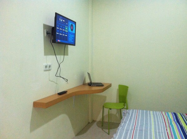 A small room with a bed, a green chair, a desk with a laptop, and a wall-mounted TV.