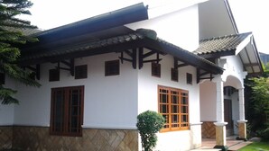 Una casa de dos plantas con tejado de baldosas, paredes blancas y base de piedra. Tiene dos ventanas con marcos de madera, un pequeño jardín con un arbusto recortado y un camino de piedra que lleva a la entrada.
